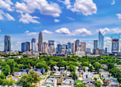 Thumbnail 13 of 13 - A cityscape with a mix of residential and commercial buildings, surrounded by lush greenery under a clear blue sky.