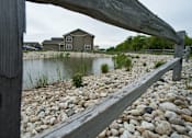 Thumbnail 3 of 13 - A wooden bench overlooks a pond with a house in the background.