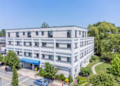 Thumbnail 12 of 12 - A modern building with a blue awning is surrounded by greenery and a clear blue sky.