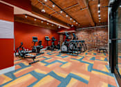 Thumbnail 6 of 15 - A gym with a variety of exercise equipment and a colorful floor at The Cordovan at Haverhill Station, Haverhill, MA