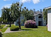 Thumbnail 5 of 17 - A house with a grey roof and a white fence is surrounded by greenery.