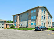 Thumbnail 25 of 25 - A blue car is parked in a parking lot in front of a large apartment building.