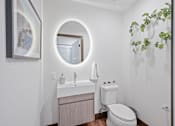 Thumbnail 15 of 49 - A bathroom with a round mirror, a sink, a toilet, and a potted plant at Sky Central Apartments, Cincinnati, 45202