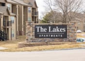 Thumbnail 16 of 16 - The Lakes apartment complex entrance sign with a stone-brick backing and the apartment buildings visible in the background.