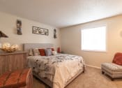 Thumbnail 17 of 17 - A spacious bedroom featuring a beige padded headboard, floral black-stitch bedding, a natural wood dresser on the left, a cozy orange lounge chair on the right, white walls, and a window offering a scenic view.