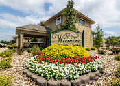 Thumbnail 1 of 17 - Exterior view of the Wilshire clubhouse, featuring a large building with a welcoming entrance, wide windows, and surrounding landscaping,