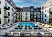 Thumbnail 1 of 30 - A swimming pool surrounded by lounge chairs in front of apartment buildings.