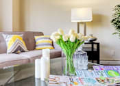 Thumbnail 4 of 16 - A living room with a grey couch, a glass table, and a vase of white flowers at Ashton Heights Apartments, Maryland, 20746