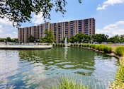 Thumbnail 38 of 46 - A large building is situated next to a body of water with a fountain in the middle.