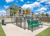 Thumbnail 41 of 44 - A green bench sits in front of a metal fence with apartment buildings in the background.