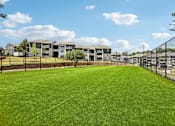 Thumbnail 6 of 40 - A large grassy field with a fence and apartment buildings in the background.