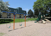 Thumbnail 25 of 30 - A playground with swings and a slide in front of a building.