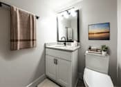 Thumbnail 10 of 18 - A bathroom with a white toilet, a sink with a marble countertop, a mirror, a towel rack, and a framed picture on the wall.