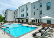 Thumbnail 17 of 21 - One Bedroom Apartments in Lakeland TN - Lakeland Town Square - Pool Area with Multiple Levels, Lounge Chairs with Cushions, white Umbrellas, Seating Area with Table, Brick Details, and Nearby Foliage