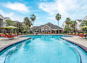 Thumbnail 1 of 16 - A large swimming pool in a residential area with red lounge chairs and umbrellas at The Fenwick Apartments, Humble, Texas
