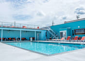 Thumbnail 17 of 22 - A large swimming pool with red lounge chairs and a blue building in the background.