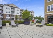 Thumbnail 15 of 29 - a large stone patio with three lounge chairs and a fire pit in front of an apartment building at Parc at Princeton Junction Apartments, New Jersey, 08540