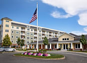 Thumbnail 1 of 35 - A large building with a flag on top and a flower bed in front at Axis at Lakeshore Apartments, Bridgewater, MA 02324