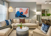 Thumbnail 16 of 28 - A living room with a beige couch, a grey coffee table, and a large abstract painting on the wall.
