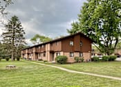 Thumbnail 1 of 11 - A long building with a brown roof and windows is surrounded by trees and grass at Root River Apartments, Greendale, WI, 53129