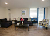 Thumbnail 19 of 28 - A living room with a black couch, a black armchair, a coffee table, and a yellow and blue patterned chair.
