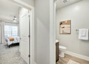 Thumbnail 11 of 37 - A white bathroom with a toilet, sink, and a bedroom with a bed and window at The Ashton at East Park Apartments, Kennesaw, Georgia