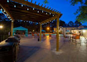 Thumbnail 1 of 23 - A patio area with a pergola and chairs is lit up at night.