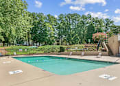 Thumbnail 10 of 26 - A large outdoor swimming pool surrounded by trees and a patio area with chairs and a table at Emerald Bay, Charlotte, NC, 28217