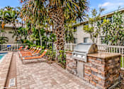 Thumbnail 34 of 35 - A patio with a grill and chairs is surrounded by a pool and trees. at Sarasota South Apartments, Florida