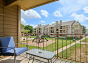 Thumbnail 15 of 39 - A balcony with a blue chair and table overlooks a grassy area and buildings.