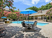 Thumbnail 33 of 39 - A poolside table with chairs and umbrellas is surrounded by a pool and trees.