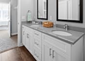 Thumbnail 6 of 34 - A bathroom with a double vanity, two mirrors, ample cabinet space, and hardwood-style flooring near a doorway to a walk-in closet and bedroom.