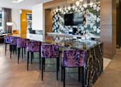 Thumbnail 19 of 34 - A community kitchen with a long bar, modern purple barstools, pendant chandelier, and a TV in a resident lounge.