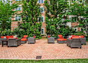 Thumbnail 35 of 59 - A patio with red cushions and grey wicker furniture is surrounded by trees and a brick building.