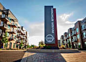 Thumbnail 26 of 34 - A River Station sign in front of modern apartment buildings with balconies and trees lining a street.