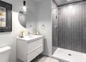 Thumbnail 30 of 39 - Steelcote Crossing bathroom with white cabinetry, white quartz countertops, large circular mirror and walk in shower with tile surround at Steelcote Square, Missouri