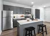 Thumbnail 20 of 39 - model kitchen with stainless steel appliances, plank flooring, kitchen island, white quartz countertops, gray cabinetry and white tile backsplash at Steelcote Square, St. Louis, 63103