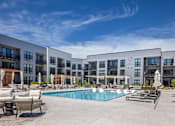 Thumbnail 2 of 37 - A large swimming pool surrounded by lounge chairs and umbrellas in front of a modern apartment complex.