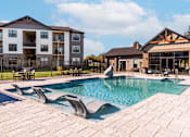 Thumbnail 6 of 18 - A large swimming pool with a patio and a building in the background.