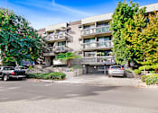 Thumbnail 9 of 12 - A black car is parked on the left side of a street in front of a building with balconies.