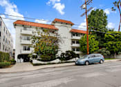 Thumbnail 1 of 4 - A blue car is parked on the street in front of a white building with a red tile roof.