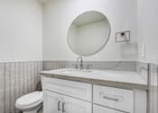 Thumbnail 16 of 21 - A white bathroom with a toilet, sink, and a round mirror.