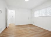 Thumbnail 8 of 11 - A room with white walls and wooden flooring with a door on the left and a window with blinds on the right.