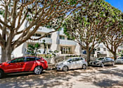 Thumbnail 9 of 11 - A red car is parked in front of a white building with trees.