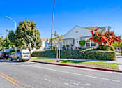 Thumbnail 9 of 11 - A street view with cars parked on the side of the road and a white house with a red flower bush in front.