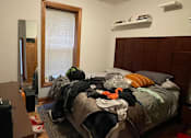 Thumbnail 10 of 13 - A bedroom with a messy bed and a window with blinds.