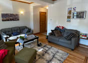 Thumbnail 3 of 13 - A living room with a grey couch, a green ottoman, a coffee table, and a ceiling fan.