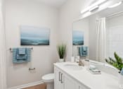 Thumbnail 27 of 41 - A bathroom with a white counter, a white toilet, a sink, a mirror, a potted plant, and a painting hanging on the wall.