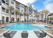 Thumbnail 2 of 13 - A swimming pool surrounded by lounge chairs and palm trees in front of a multi-story apartment building at The Addison, Baton Rouge, LA
