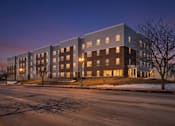 Thumbnail 14 of 18 - A large, modern building with a flat roof and a mix of dark and light brown windows is illuminated from the inside by lights.